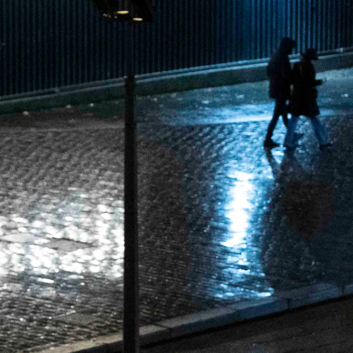 Jean-Paul · 20/12/2025 · 1623 x 1623 Cette photographie capture deux silhouettes marchant sous la pluie nocturne. Le **sujet principal** est l’interaction entre les personnes et leur environnement humide.La **composition** est symétrique, avec une perspective reflétée sur une surface mouillée. Le **cadrage** est serré, centré sur les personnages et leur environnement immédiat.La **lumière** est tamisée, avec des éclairages artificiels diffusant une ambiance bleutée. Les **couleurs dominantes** sont les tons gris et bleus, renforçant une atmosphère froide et humide.Le **style** est réaliste et légèrement flou, créant une **ambiance** mélancolique et contemplative.