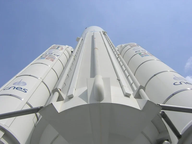 Frédéric · 23/08/2006 · 1600 x 1200 Cette photographie met en scène un lanceur spatial Ariane, sujet principal imposant et central. La composition montre le bas du lanceur avec ses boosters latéraux, capturés depuis une perspective basse, accentuant sa hauteur et sa puissance.Le cadrage est serré, mettant en valeur les détails techniques. La lumière est naturelle et claire, soulignant les contours métalliques. Les couleurs dominantes sont le gris métallique et le bleu pâle du ciel, créant un contraste sobre et professionnel. Le style est documentaire et technique, évoquant une ambiance industrielle et scientifique, mêlant innovation et rigueur.