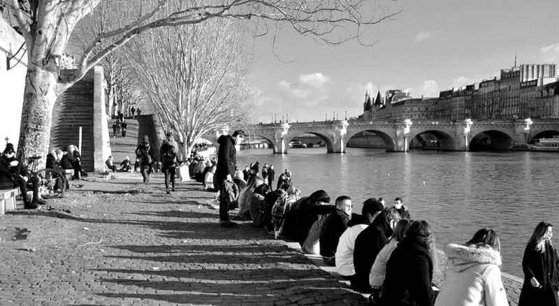 Pascale · 09/01/2021 · 1000 x 548 Cette photographie en noir et blanc capture une scène urbaine le long d'une rivière, probablement la Seine. Le sujet principal est un groupe de personnes assises sur une berge, observant le paysage. La composition est équilibrée, avec la rivière au centre et des ponts en arrière-plan.Le cadrage est large, incluant des arbres dénudés et des bâtiments. La lumière est douce, suggérant un moment en fin de journée. Les tons gris dominent, accentuant les contrastes entre ombres et lumières. Le style est documentaire et réaliste, évoquant une ambiance paisible et contemplative.