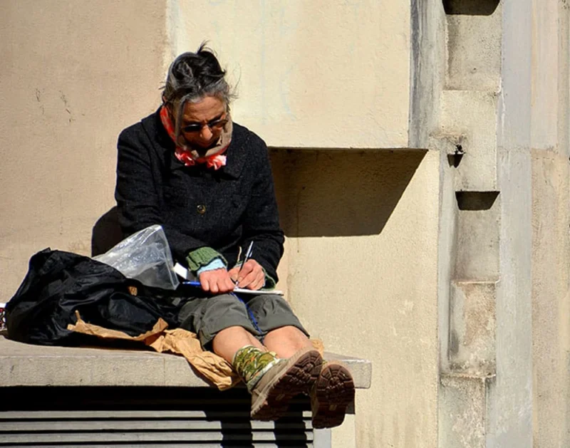 Pascale · 04/04/2020 · 1000 x 786 Cette photographie montre une personne assise sur un banc, semblant manger un sandwich. Le sujet principal est cette personne, vêtue d’un manteau sombre et d’un bonnet, avec un sac à ses côtés.La composition est simple et naturelle, avec un cadrage serré mettant en valeur le sujet. La lumière est douce et naturelle, probablement en journée, créant des ombres légères sur le mur et le banc.Les couleurs dominantes sont les tons neutres (beige, gris) avec des touches de rouge et de vert. Le style est réaliste et documentaire, capturant un moment de vie quotidienne. L’ambiance est calme et intimiste.