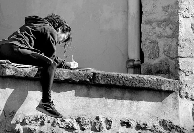 Pascale · 22/03/2026 · 1000 x 682 Cette photographie en noir et blanc met en scène un homme assis, sujet principal, penché en avant comme absorbé par une activité (peinture ou dessin). La composition est simple et épurée, avec un cadrage serré sur le sujet et son environnement immédiat.La lumière naturelle, douce et contrastée, accentue les ombres et les textures. Les couleurs dominantes, bien que monochromes, jouent sur les nuances de gris, renforçant l’ambiance contemplative et mélancolique. Le style est réaliste et documentaire, évoquant une scène du quotidien. L’ambiance est calme et introspective, presque poétique.