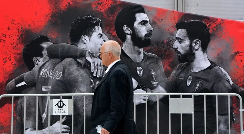 Pascale · 12/06/2018 · 1000 x 548 Cette photographie met en scène un groupe de supporters de football devant une grande affiche murale. Le sujet principal est un homme en premier plan, portant un sweat à capuche, observant les joueurs représentés sur le mural.La composition joue sur le contraste entre le supporter réel et les images stylisées des joueurs. Le cadrage est serré sur le groupe et l’affiche, créant une ambiance immersive. Les couleurs dominantes sont le rouge, le noir et le gris, renforçant une ambiance intense et passionnée. Le style est réaliste pour les visages et graphique pour le fond, évoquant une énergie vibrante et une dévotion sportive.