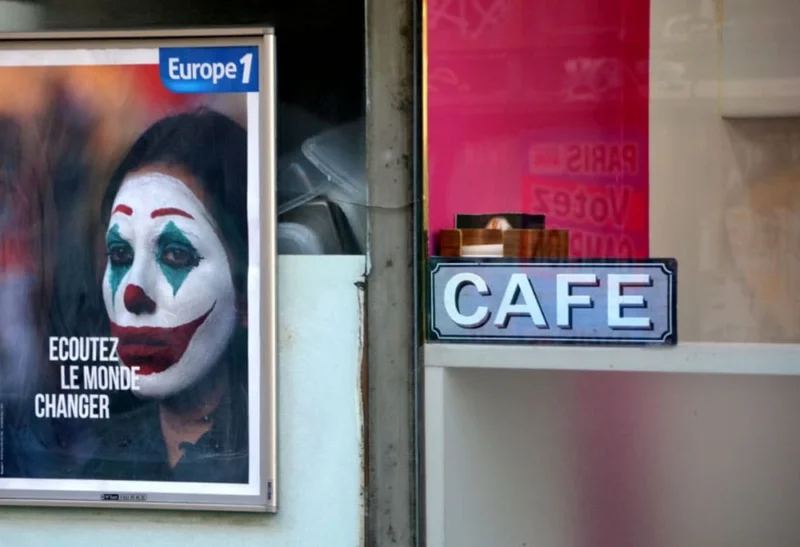 Pascale · 25/03/2020 · 1000 x 684 Cette photographie met en scène une affiche de journal "Europe 1" représentant un clown, avec le texte "Écoutez le monde changer". À côté, une enseigne lumineuse "CAFÉ" est partiellement visible. La composition juxtapose ces deux éléments, créant un contraste entre le visuel inquiétant du clown et l'ambiance plus chaleureuse du café.Le cadrage est serré, avec une lumière naturelle douce et des couleurs dominées par des tons froids (bleu, gris) et chauds (rose, rouge). L'ambiance est à la fois mystérieuse et urbaine, mêlant une touche de mélancolie et de vie quotidienne.