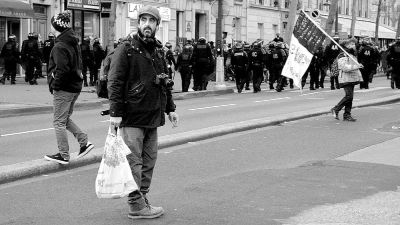 Pascale · 11/01/2020 · 1000 x 564 Cette photographie en noir et blanc montre une manifestation. Le sujet principal est un homme en premier plan, marchant avec une veste épaisse et une écharpe, tenant un sac plastique. La composition met en valeur la foule en arrière-plan, avec des personnes tenant des pancartes. Le cadrage est serré sur l’homme, tout en incluant une partie de la rue et des manifestants. La lumière est naturelle, sans contraste marqué, créant une ambiance sobre et réaliste. Les tons gris dominent, renforçant une impression de sérieux et de gravité. Le style est documentaire, captant un moment authentique. L’ambiance est calme mais engagée.