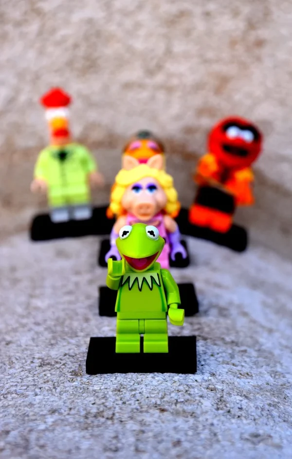 Franck TREBOIT · 14/03/2026 · 892 x 1400 Cette photographie met en scène quatre figurines Lego inspirées de personnages de Muppets. Le sujet principal est la figurine verte en avant-plan, assise dans une voiture miniature. La composition est symétrique et alignée, avec les personnages disposés en ligne derrière elle. Le cadrage est serré, mettant l'accent sur les détails des figurines. La lumière est douce et naturelle, éclairant uniformément la scène. Les couleurs dominantes sont vives et variées : vert, orange, jaune et rouge. Le style est ludique et coloré, évoquant une ambiance joyeuse et enfantine.