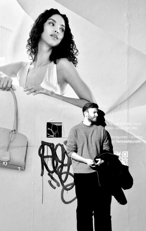 Franck TREBOIT · 02/03/2026 · 886 x 1400 Cette photographie en noir et blanc met en scène deux sujets principaux : une femme en gros plan sur un écran mural et un homme en bas, de dos, tenant un sac.La composition joue sur les contrastes entre le fond géométrique et les formes organiques. Le cadrage est serré sur l’homme, tandis que la femme semble flotter derrière lui.La lumière est douce, créant des ombres nettes. Les couleurs dominantes, bien que monochromes, exploitent les contrastes de tons clairs et foncés.Le style est artistique et moderne, évoquant une ambiance à la fois intime et surréaliste.