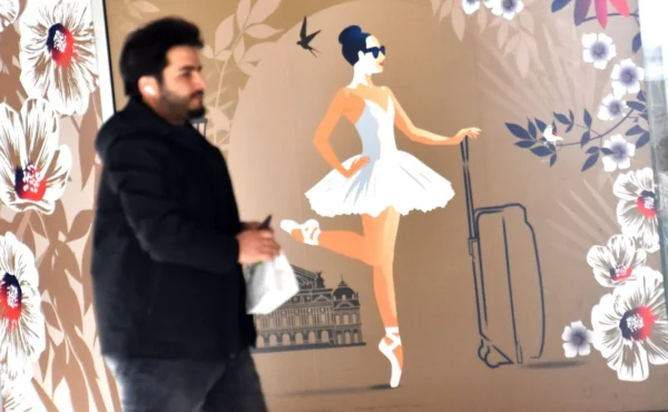 Franck TREBOIT · 07/03/2026 · 1400 x 864 Cette photographie montre un homme marchant devant une fresque murale. Le sujet principal est cette illustration stylisée représentant une ballerine en tutu tenant une valise à roulettes.La composition juxtapose l’homme réel et l’image onirique. Le cadrage est serré sur l’homme, avec la fresque en arrière-plan. La lumière semble naturelle, douce et diffuse. Les couleurs dominantes sont des tons pastel (beige, rose pâle) avec des touches de bleu et de vert.Le style est artistique et graphique, évoquant une ambiance à la fois rêveuse et légèrement rétro. L’ensemble dégage une impression de légèreté et de fantaisie.