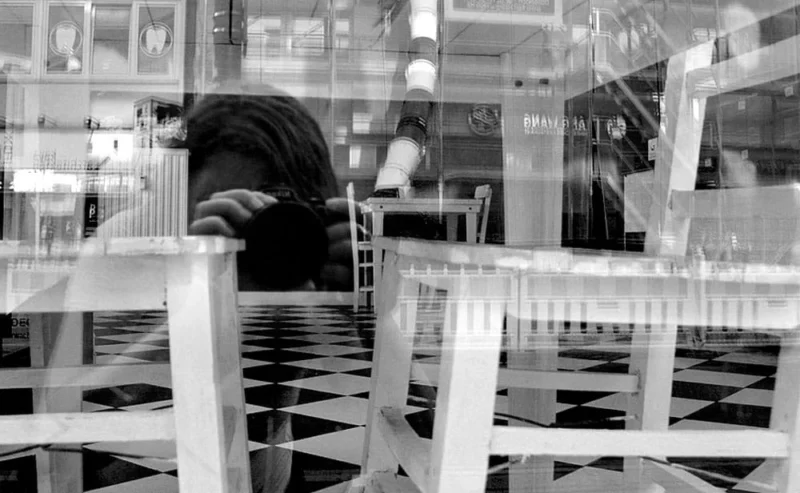Pascale · 15/07/2022 · 1000 x 616 Cette photographie en noir et blanc met en scène un espace minimaliste et architectural. Le sujet principal semble être une personne assise, partiellement cachée, dans un lieu rempli de tables et chaises blanches disposées de manière ordonnée. La composition joue sur les lignes droites et la symétrie, renforçant une ambiance épurée et géométrique.Le cadrage est serré, centré sur les éléments architecturaux et les jeux d'ombres. La lumière, douce et diffuse, accentue les contrastes entre clarté et obscurité, créant une atmosphère à la fois moderne et mystérieuse. Les couleurs dominantes, bien que monochromes, exploitent les nuances de gris et de blanc pour souligner la pureté et la simplicité du style.