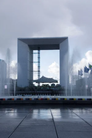 Pascale · 28/03/2026 · 1167 x 1750 Cette photographie met en scène une grande structure architecturale en forme d'arc, entourée de jets d'eau. Le sujet principal est cette arche transparente, encadrée par des jets d’eau qui créent une brume légère.La composition est symétrique et centrée, avec un cadrage serré sur l’arche. La lumière, diffuse et tamisée, donne une ambiance mystérieuse et légèrement dramatique. Les couleurs dominantes sont les gris et bleus froids, adoucis par les nuances blanches de la brume.Le style est épuré et contemporain, évoquant une ambiance futuriste et calme.