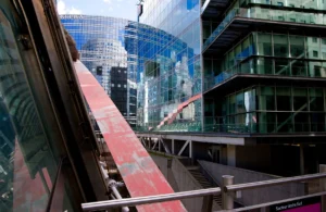 Pascale · 28/03/2026 · 1750 x 1137 Cette photographie met en scène un bâtiment moderne avec une architecture futuriste. Le sujet principal est la structure en verre et acier, reflétant des teintes bleutées et des éléments urbains.La composition joue sur les lignes diagonales et les perspectives, accentuées par le cadrage serré et les balustrades métalliques. La lumière naturelle éclaire la scène, créant des contrastes entre ombres et reflets.Les couleurs dominantes sont les bleus, gris et touches de rouge, évoquant un style contemporain et dynamique. L’ambiance est à la fois urbaine et innovante, avec une impression de mouvement et de modernité.