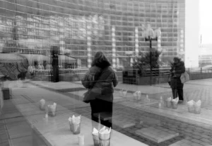 Pascale · 01/04/2026 · 1750 x 1199 Cette photographie en noir et blanc met en scène deux personnes en mouvement, floutées, dans un espace urbain. Le sujet principal semble être une femme penchée, entourée de petites bougies allumées sur le sol.La composition joue sur les contrastes entre les formes floues et les éléments nets, comme les bougies et les bâtiments en arrière-plan. Le cadrage est serré sur les personnages et les bougies, avec une profondeur de champ réduite.La lumière est douce, créant des ombres marquées. Les couleurs dominantes sont les tons de gris, typiques du noir et blanc, renforçant une ambiance mélancolique et contemplative. Le style est documentaire et poétique.