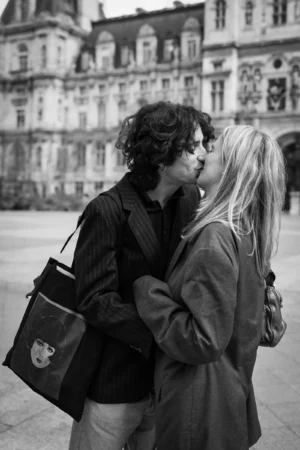 Claude · 3234 x 4851 Cette photographie met en scène un couple en train de s'embrasser tendrement. Le sujet principal est leur complicité, captée de manière intime.La composition est simple et centrée sur eux, avec un cadrage serré qui isole les deux personnages. L'arrière-plan, flou, montre un bâtiment classique, suggérant un cadre urbain historique.La lumière est douce, naturelle, et semble provenir d'une source latérale, créant des contrastes subtils. En noir et blanc, les tons varient du clair au foncé, accentuant les textures des vêtements et des visages.Le style est documentaire et romantique, évoquant une ambiance chaleureuse et nostalgique.