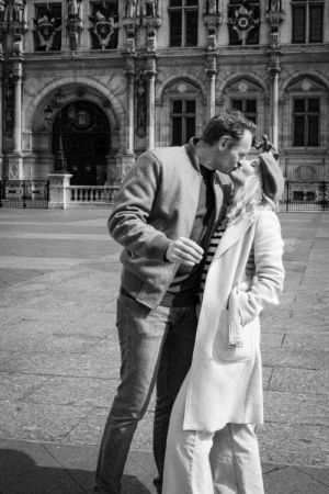 Claude · 3090 x 4635 Cette photographie en noir et blanc met en scène un couple en extérieur. Le sujet principal est un homme et une femme en étreinte tendre, l’homme ajustant le col de la veste de la femme.La composition est centrée sur eux, avec un cadrage serré qui isole leur interaction. L’arrière-plan architectural, avec ses colonnes et détails classiques, ajoute une touche d’élégance et de profondeur.La lumière douce, probablement naturelle, crée des contrastes subtils. Les tons dominants, en niveaux de gris, renforcent l’ambiance romantique et intemporelle, évoquant une scène à la fois intime et poétique.