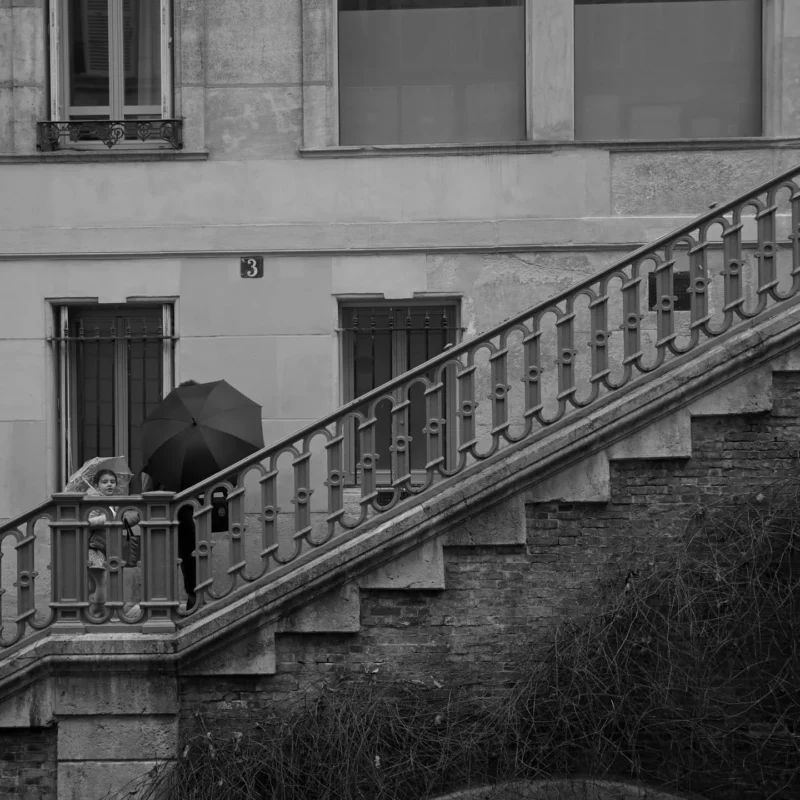 Jojo · 1440 x 1440 Cette photographie en noir et blanc met en scène un escalier extérieur en pierre avec une rampe en fer forgé. Le sujet principal est une personne, partiellement visible, assise en bas des marches avec un sac à dos.La composition repose sur une perspective ascendante, guidant le regard vers le bâtiment en arrière-plan. Le cadrage est serré, accentuant les détails architecturaux et l’ombre. La lumière, douce et naturelle, crée des contrastes marqués entre clairs et ombres, renforçant l’ambiance mélancolique et contemplative. Les tons gris dominent, soulignant une atmosphère calme et introspective. Le style est documentaire et poétique.