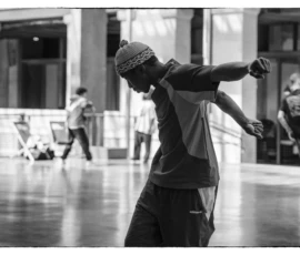 Thierry · 22/04/2026 · 3840 x 2560 Cette photographie en noir et blanc met en scène un danseur ou skateur comme sujet principal, en mouvement dynamique, bras tendu. La composition est centrée sur lui, avec des éléments d’arrière-plan flous, comme des personnes et des structures architecturales.Le cadrage est serré, capturant l’énergie du sujet. La lumière est naturelle, contrastée, créant des ombres marquées. L’ambiance est urbaine et spontanée, évoquant un moment de performance ou de pratique dans un espace public. Le style est documentaire et artistique, mettant l’accent sur le mouvement et la texture.