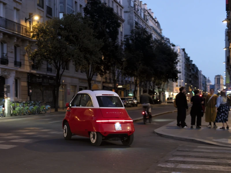 Cette photographie met en scène une **voiture rouge compacte** (une Fiat 500) comme sujet principal. La **composition** montre une rue urbaine animée avec des bâtiments en arrière-plan et des passants.Le **cadrage** est serré sur la voiture, mais inclut aussi une partie du trottoir et des arbres. La **lumière** est douce, typique d’un **crépuscule**, créant une ambiance tamisée et chaleureuse. Les **couleurs dominantes** sont les tons rouges de la voiture, les gris des bâtiments et les verts des arbres.Le style est **réaliste et documentaire**, évoquant une scène de vie quotidienne en ville, avec une **ambiance paisible et légèrement nostalgique**.