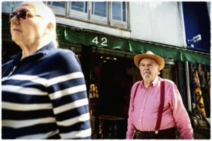 Thierry · 25/04/2026 · 3840 x 2560 Cette photographie met en scène deux hommes âgés devant une porte numérotée 42. Le sujet principal semble être l’homme à droite, vêtu d’un chapeau et d’une chemise rose, observant la scène. La composition joue sur le contraste entre les deux personnages : l’un en mouvement (strié), l’autre statique.Le cadrage est serré, centré sur leurs visages et torses. La lumière est naturelle, probablement en extérieur, avec des ombres marquées. Les couleurs dominantes sont sobres : bleu et blanc pour le premier homme, rose et beige pour le second, avec des tons neutres en arrière-plan.L’ambiance est à la fois réaliste et légèrement mélancolique, évoquant une scène du quotidien.