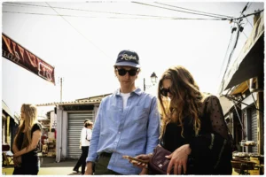 Thierry · 25/04/2026 · 3840 x 2560 Cette photographie met en scène deux jeunes comme sujets principaux, un homme en chemise bleue et chapeau, et une femme aux cheveux longs. Le cadrage est serré, centré sur eux, avec un arrière-plan flou montrant des éléments urbains et des personnes.La lumière est douce, légèrement surexposée, créant une ambiance chaleureuse et naturelle. Les couleurs dominantes sont des tons neutres (bleu, gris) avec des touches de brun et de blanc. Le style est décontracté et spontané, évoquant une scène de rue ou un événement informel. L’ambiance est détendue et légèrement nostalgique.