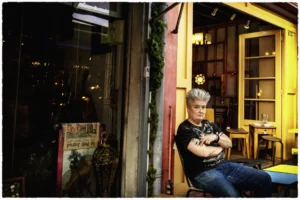 Thierry · 25/04/2026 · 3840 x 2560 Cette photographie met en scène un homme assis sur une moto devant une boutique. Le sujet principal est cet individu, vêtu d’une veste en cuir, avec une attitude décontractée et un peu mélancolique.La composition joue sur le contraste entre l’intérieur éclairé de la boutique et l’extérieur plus sombre. Le cadrage est serré, centré sur le sujet avec un arrière-plan légèrement flou. La lumière est tamisée, créant une ambiance intimiste et nocturne.Les couleurs dominantes sont des tons chauds (bois, jaune) et des teintes sombres (noir, gris), renforçant une atmosphère rétro et urbaine. Le style rappelle le photojournalisme documentaire.
