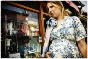 Thierry · 25/04/2026 · 3840 x 2560 Cette photographie met en scène une femme enceinte, sujet principal, vêtue d’une robe à motifs floraux. Le cadrage est serré sur elle, avec un arrière-plan flou montrant une boutique artisanale colorée.La lumière semble naturelle, avec des contrastes doux, mettant en valeur les détails de sa tenue et son visage. Les couleurs dominantes sont vives et chaudes : tons pastel de la robe, teintes terreuses et éclatantes des objets en arrière-plan.Le style est documentaire et spontané, évoquant une ambiance authentique et chaleureuse, presque intimiste malgré la présence du commerce en toile de fond.