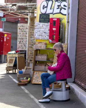 Thibault · 25/04/2026 · 1535 x 1920 Cette photographie montre une femme âgée, sujet principal, assise sur un tabouret devant un magasin rempli de téléphones publics rouges. La composition met en valeur son visage et ses mains occupées par un appareil photo.Le cadrage est serré, centré sur elle, avec des éléments urbains en arrière-plan. La lumière semble naturelle, légèrement diffuse, créant des ombres douces. Les couleurs dominantes sont les tons froids (gris, bleu) et chauds (rouge des téléphones, violet de son vêtement).Le style est documentaire, capturant un instant de vie quotidienne. L’ambiance est calme et nostalgique, évoquant une époque révolue.
