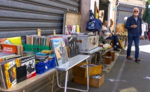 Thibault · 25/04/2026 · 1920 x 1186 Cette photographie montre un **stand de livres d’occasion** en extérieur, probablement un marché. Le **sujet principal** est l’étalage de livres variés, bien fournis et disposés sur des tables en bois.La **composition** est serrée, avec une perspective légèrement basse, mettant en valeur les livres et les objets autour. Le **cadrage** inclut aussi un homme en arrière-plan, ajoutant une touche de vie quotidienne. La **lumière** est naturelle, douce, sans ombre marquée, suggérant un jour nuageux.Les **couleurs dominantes** sont neutres (beige, gris) avec des touches vives (jaunes, rouges) sur les couvertures. L’ambiance est **rustique et chaleureuse**, évoquant un lieu convivial et authentique.
