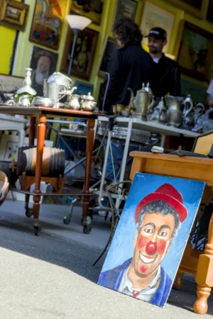 Thibault · 25/04/2026 · 1280 x 1920 Cette photographie montre un stand de marché comme sujet principal. La composition met en avant une peinture colorée représentant un clown au sourire exagéré, en premier plan. Le cadrage inclut aussi des objets métalliques anciens en arrière-plan, ainsi que deux personnes floues.La lumière semble naturelle, probablement ensoleillée, créant des contrastes nets. Les couleurs dominantes sont vives : bleu, rouge et jaune pour la peinture, et des tons métalliques froids pour les objets.Le style est réaliste et spontané, évoquant une ambiance de marché artisanal et chaleureuse.