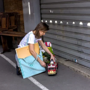 Thibault · 25/04/2026 · 1920 x 1920 Cette photographie montre une jeune fille, sujet principal, assise sur le sol près d’un trottoir. Elle semble organiser des objets dans un sac à dos bleu.La composition est simple et naturelle, avec un cadrage serré sur la scène. La lumière est claire et naturelle, probablement en journée. Les couleurs dominantes sont les teintes pastel (bleu, beige) et des touches de rouge et vert.Le style est documentaire et spontané, capturant un moment du quotidien. L’ambiance est calme et réaliste, évoquant une activité simple et ordinaire.