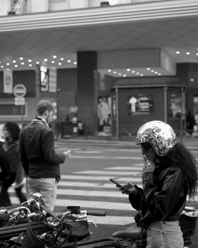 Jojo · 11/04/2026 · 3072 x 3840 Cette photographie en noir et blanc met en scène un homme en tenue de motard, casque à la main, absorbé par son téléphone portable. Le sujet principal est donc cet individu, isolé au premier plan.La composition joue sur le contraste entre lui et le fond flou, où des personnes et des éléments urbains (bâtiments, enseignes) suggèrent un lieu public, probablement une gare. Le cadrage serré sur le motard crée une impression d’intimité malgré le cadre urbain.La lumière est douce, sans ombres marquées, renforçant une ambiance moderne et neutre. Le style est documentaire, réaliste, avec une touche d’introspection. L’absence de couleurs accentue les contrastes et les textures.