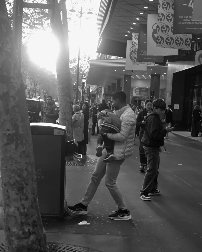 Jojo · 11/04/2026 · 3072 x 3840 Cette photographie en noir et blanc capture une scène urbaine animée. Le sujet principal est un homme en mouvement, vêtu d’un sweat et de baskets, qui semble marcher rapidement. La composition montre une foule en arrière-plan, sous des enseignes et des bâtiments.Le cadrage est serré sur le sujet avant-plan, avec une profondeur de champ qui floute légèrement les personnes derrière. La lumière, vive et contrastée, provient probablement du soleil bas, créant des ombres marquées. L’ambiance est dynamique et quotidienne, évoquant une ville active. Le style est documentaire et spontané, renforcé par le traitement monochrome.