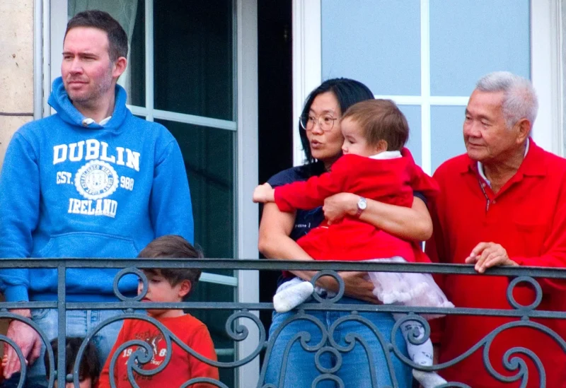 Pascale · 01/03/2026 · 1500 x 1029 Cette photographie montre une famille sur un balcon. Le sujet principal est un homme âgé tenant un enfant, entouré d’une femme et d’un autre homme en bleu portant un sweat "Dublin Ireland".La composition est serrée, avec un cadrage en plan moyen. La lumière est naturelle et douce, probablement en journée. Les couleurs dominantes sont les tons chauds : rouge (vêtements), bleu (sweat) et gris (balcon et murs).Le style est documentaire et spontané, capturant un moment familial. L’ambiance est chaleureuse et conviviale.