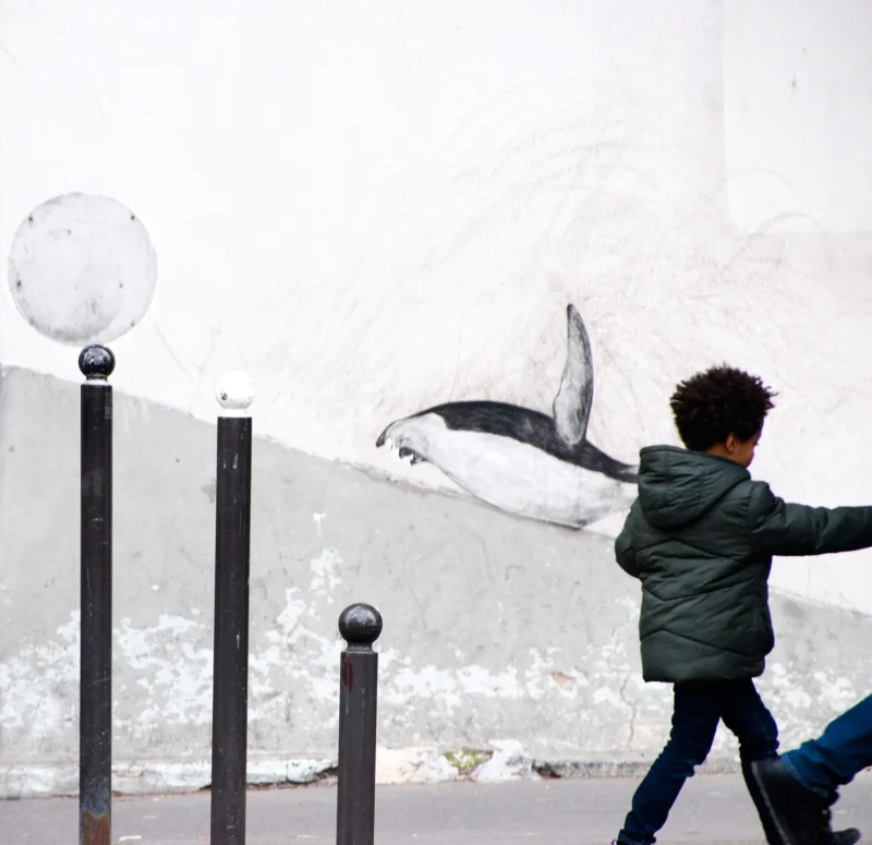 Pascale · 01/03/2026 · 1750 x 1696 Cette photographie met en scène un enfant comme sujet principal, marchant devant un mur tagué représentant un poisson. La composition joue sur le contraste entre le mouvement du garçon et l'immobilité du graffiti. Le cadrage est serré, centré sur l’enfant et les poteaux noirs en premier plan, créant une perspective dynamique.La lumière est naturelle et diffuse, adoucissant les contours. Les couleurs dominantes sont les tons gris (mur, vêtements) et noir (poteaux), avec une touche de bleu et de blanc dans le graffiti. Le style est réaliste et urbain, évoquant une ambiance à la fois quotidienne et artistique.