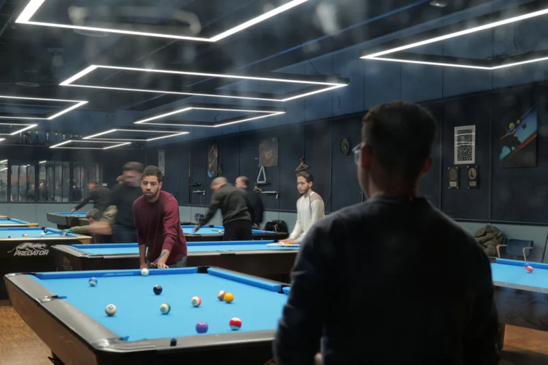 Bao · 29/12/2025 · 5900 x 3933 Cette photographie montre un groupe jouant au billard dans un bar. Le sujet principal est un homme vu de dos, observant la partie. La composition met en scène plusieurs tables de billard et des joueurs en arrière-plan. Le cadrage est serré sur l’action, avec une profondeur de champ moyenne.La lumière est douce et artificielle, éclairant uniformément la scène. Les couleurs dominantes sont les bleus (tables) et les tons chauds (vêtements, éclairage). Le style est réaliste et contemporain, évoquant une ambiance conviviale et détendue, typique d’un lieu de loisirs.