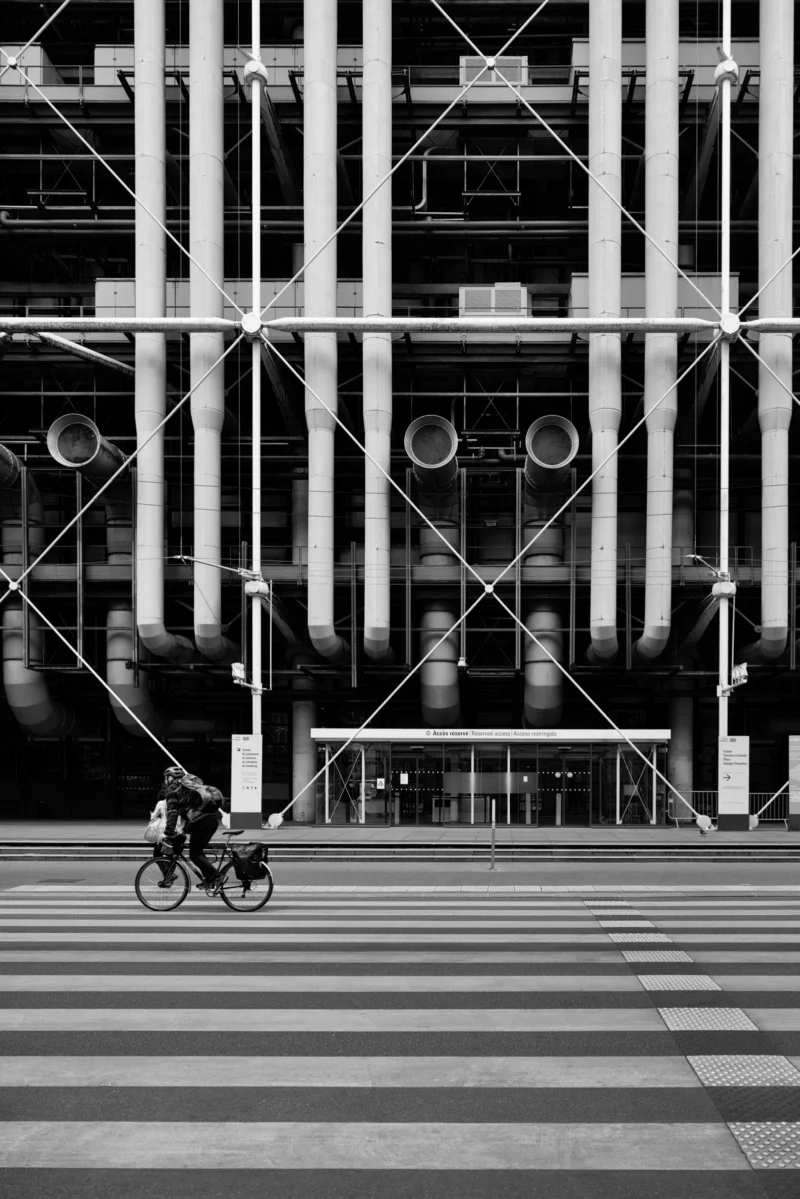 Philippe · 31/03/2026 · 2668 x 4000 Cette photographie en noir et blanc met en scène un cycliste comme sujet principal, traversant une rue en damier. La composition joue sur les lignes géométriques des bâtiments et des pavés, créant un effet de perspective et de symétrie. Le cadrage serré et bas accentue la profondeur et l’architecture imposante.La lumière, contrastée, souligne les ombres et les formes architecturales. Les couleurs dominantes, bien que monochromes, exploitent les nuances de gris pour renforcer le contraste. Le style est épuré et urbain, évoquant une ambiance moderne et dynamique, presque cinématographique.