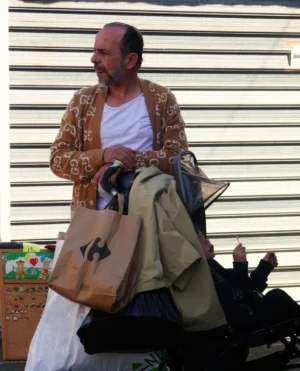 Bao · 2520 x 3113 Cette photographie montre un homme comme sujet principal, vêtu d’un gilet coloré et d’un t-shirt blanc, tenant des sacs. La composition met en avant un cadrage serré sur lui, avec des éléments en arrière-plan flous (table, objets divers).La lumière semble naturelle, probablement en extérieur, avec des ombres douces. Les couleurs dominantes sont les tons chauds du gilet, contrastant avec les teintes neutres des vêtements et du fond.Le style est documentaire et spontané, évoquant une ambiance de marché ou de rue, avec une touche de simplicité et d’authenticité.