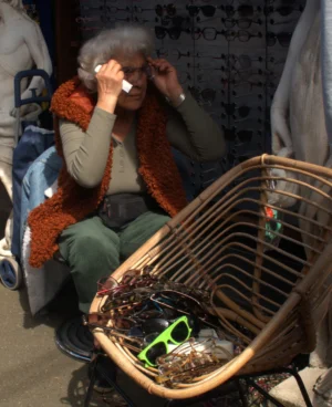 Bao · 2798 x 3436 Cette photographie montre une personne âgée, sujet principal, assise et tenant un panier en osier rempli de déchets ou d’objets divers. Le cadrage est serré, mettant en valeur son visage et le panier. La lumière semble naturelle mais tamisée, créant des ombres douces.Les couleurs dominantes sont des tons neutres (beige, gris) avec des touches de vert et de rouge (vêtements). Le style est documentaire, capturant un moment réaliste et spontané. L’ambiance est sobre et un peu mélancolique, évoquant une scène de vie quotidienne ou sociale.
