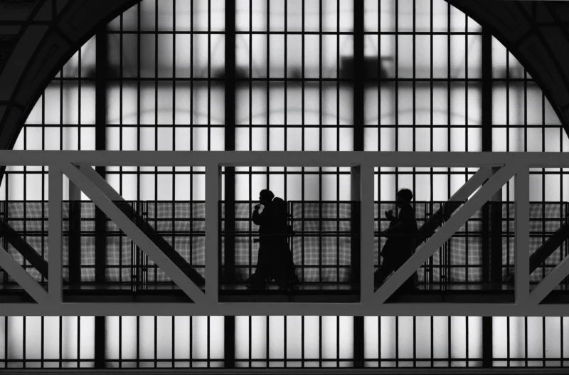 Philippe · 13/11/2025 · 3000 x 1978 Cette photographie en noir et blanc met en scène deux silhouettes assises sur une structure métallique, probablement un pont ou une passerelle. Le sujet principal est ces deux personnes, presque fusionnées dans l’ombre.La composition repose sur une architecture industrielle avec des arcs et des vitraux, créant un cadrage symétrique et géométrique. La lumière, tamisée et contrastée, accentue les ombres et les formes, renforçant une ambiance mélancolique et contemplative. Les tons dominants sont les noirs profonds et les gris, typiques du style photographique noir et blanc, évoquant une atmosphère à la fois mystérieuse et intemporelle.