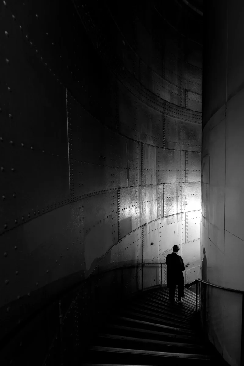 Philippe · 30/11/2025 · 2001 x 3000 Cette photographie en noir et blanc met en scène un homme seul, silhouette solitaire, descendant un escalier éclairé. Le sujet principal est ce personnage, isolé dans un espace architectural moderne aux lignes épurées.La composition joue sur les contrastes entre lumière et ombre, avec un éclairage directionnel qui accentue la profondeur. Le cadrage serré et la perspective des marches guident le regard vers le bas.L’ambiance est mystérieuse et contemplative, renforcée par les tons sombres et la nuit étoilée en arrière-plan. Le style est sobre, presque cinématographique, évoquant solitude et introspection.