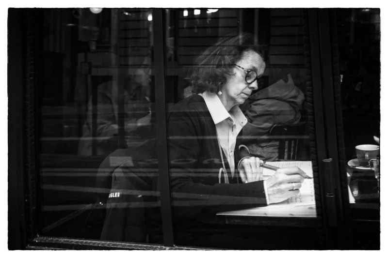 Thierry · 11/04/2026 · 3840 x 2560 Cette photographie en noir et blanc met en scène une personne âgée, sujet principal, assise à un bureau et lisant ou écrivant. La composition est simple et centrée, avec un cadrage serré sur le visage et les mains.La lumière est douce et tamisée, créant des contrastes subtils. Les couleurs dominantes, bien que limitées au noir et blanc, suggèrent des tons clairs et sombres pour les détails.Le style est documentaire et intime, évoquant une ambiance calme et réfléchie, presque mélancolique. L'image dégage une sensation de solitude et de concentration.