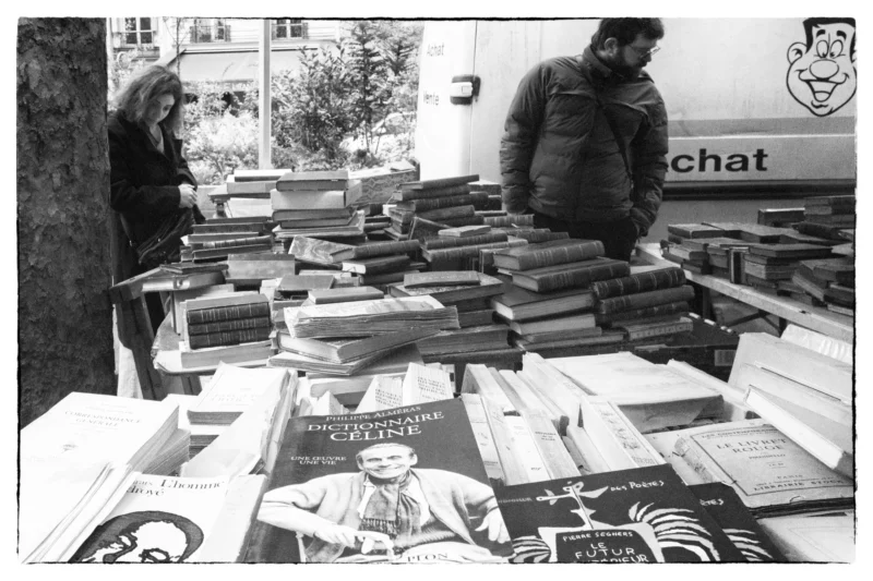Thierry · 11/04/2026 · 3840 x 2560 Cette photographie en noir et blanc montre deux personnes dans une librairie ou un stand de livres d’occasion. Le sujet principal est la multitude de livres entassés sur des tables et au sol, créant une ambiance dense et désordonnée.La composition est dynamique, avec un cadrage serré sur les livres et les personnes en arrière-plan. La lumière semble naturelle, douce et diffuse, sans ombres marquées. Les couleurs dominantes sont les nuances de gris, typiques du noir et blanc, renforçant une ambiance rétro ou vintage. Le style est documentaire, capturant un moment authentique dans un espace chaleureux et studieux.