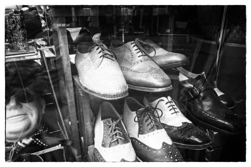 Thierry · 11/04/2026 · 3840 x 2560 Cette photographie montre principalement une vitrine remplie de chaussures masculines, principalement des modèles classiques comme des derbies et des brogues. La composition est serrée, mettant en valeur les détails des chaussures disposées sur des présentoirs métalliques.Le cadrage est proche, offrant une vue détaillée. La lumière est tamisée, créant des contrastes nets entre les zones éclairées et ombragées. En noir et blanc, les couleurs dominantes sont absentes, mais les nuances de gris renforcent les textures et les formes.Le style est intemporel et élégant, évoquant une ambiance rétro, typique d’un magasin traditionnel de chaussures haut de gamme.