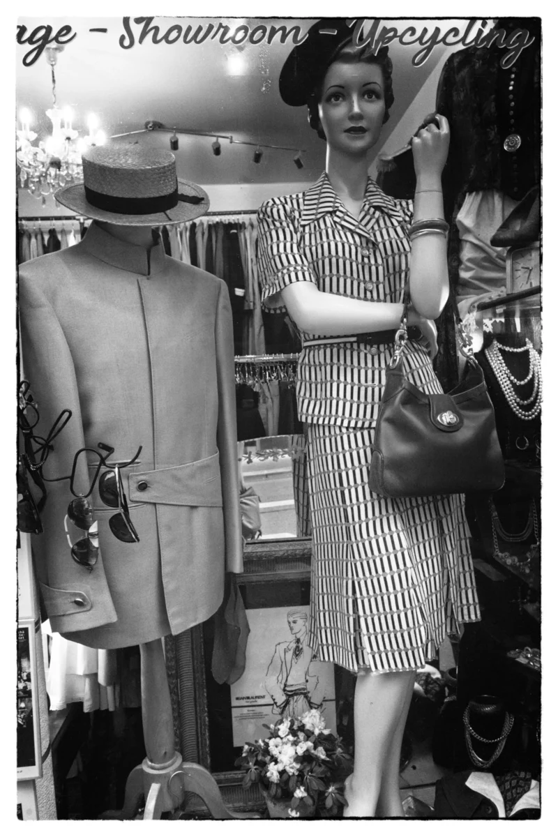Thierry · 11/04/2026 · 2560 x 3840 Cette photographie en noir et blanc montre une vitrine de magasin vintage. Le sujet principal est une mannequin vêtue d’une robe à rayures et d’un chapeau élégant, posant devant des mannequins habillés de tenues rétro (vestes, chapeaux). La composition est symétrique, avec un cadrage serré sur les mannequins et accessoires.La lumière est douce, mettant en valeur les détails. Les couleurs, bien que monochromes, évoquent des tons gris et noirs, renforçant l’ambiance rétro. Le style rappelle les années 1950-1960, créant une ambiance chic et nostalgique.