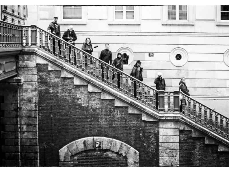 Thierry · 11/04/2026 · 3840 x 2560 Cette photographie en noir et blanc montre un groupe de personnes descendant un escalier extérieur en pierre, bordé de balustrades métalliques. Le sujet principal est l’interaction sociale et le mouvement collectif.La composition met en valeur la perspective ascendante, renforcée par le cadrage serré sur les marches. La lumière, douce et uniforme, crée des contrastes modérés, accentuant les textures architecturales. Les tons dominants, gris et noirs, renforcent une ambiance à la fois classique et mélancolique. Le style, sobre et épuré, évoque une esthétique documentaire, tandis que l’ambiance, à la fois dynamique et contemplative, suggère une scène du quotidien teintée de nostalgie.