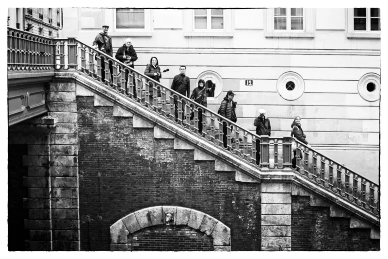 Thierry · 11/04/2026 · 3840 x 2560 Cette photographie en noir et blanc montre un groupe de personnes descendant un escalier extérieur en pierre, bordé de balustrades métalliques. Le sujet principal est l’interaction sociale et le mouvement collectif.La composition met en valeur la perspective ascendante, renforcée par le cadrage serré sur les marches. La lumière, douce et uniforme, crée des contrastes modérés, accentuant les textures architecturales. Les tons dominants, gris et noirs, renforcent une ambiance à la fois classique et mélancolique. Le style, sobre et épuré, évoque une esthétique documentaire, tandis que l’ambiance, à la fois dynamique et contemplative, suggère une scène du quotidien teintée de nostalgie.