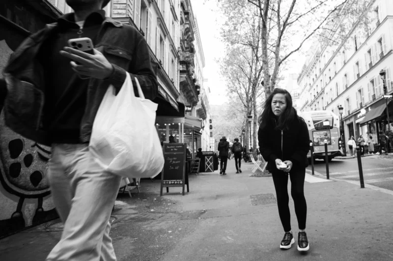 Alice · 11/04/2026 · 4820 x 3213 Cette photographie en noir et blanc met en scène une jeune femme comme sujet principal, marchant sur un trottoir urbain. La composition joue sur les contrastes entre ombre et lumière, avec un cadrage serré sur elle en arrière-plan, tandis qu’un individu avec un sac à dos coloré domine l’avant-plan.La lumière est naturelle, probablement matinale ou vesprée, créant des jeux d’ombres marquées. Les couleurs, bien que monochromes, suggèrent des tons gris et noirs, avec des touches de blanc.Le style est documentaire et spontané, évoquant une ambiance urbaine authentique et mélancolique.