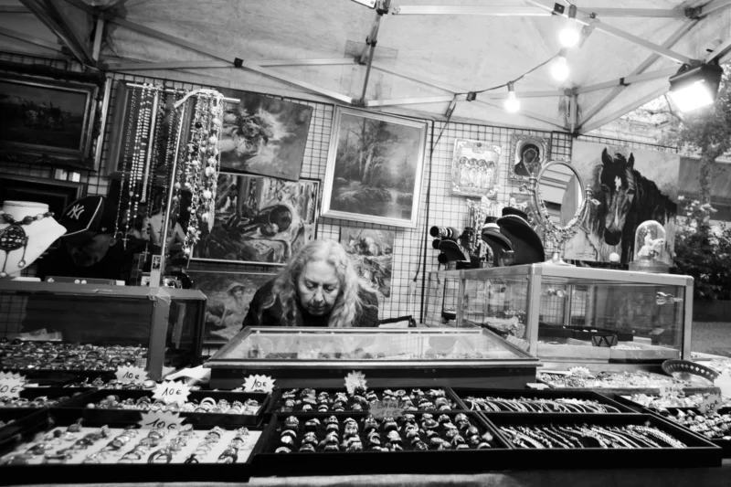 Alice · 11/04/2026 · 4445 x 2963 Cette photographie en noir et blanc montre un **marché aux puces ou une boutique d'antiquités**. Le **sujet principal** est un homme assis derrière un étalage rempli d’objets anciens (montres, bijoux, instruments).La **composition** est dense, avec des étagères chargées et un cadrage serré. La **lumière** est douce mais contrastée, mettant en valeur les détails. L’**ambiance** est rétro et nostalgique, renforcée par le style **monochrome**.Les **couleurs dominantes** sont absentes (noir et blanc), accentuant l’aspect vintage. L’atmosphère évoque un lieu chargé d’histoire et de curiosités.
