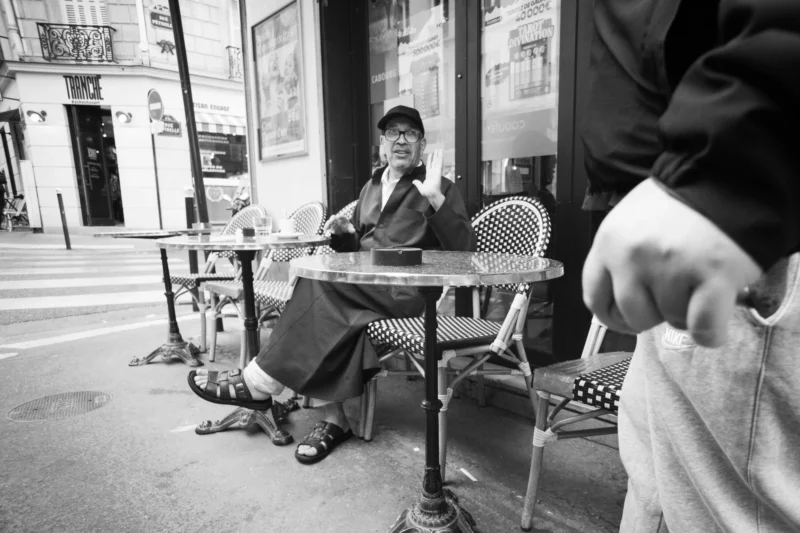 Alice · 11/04/2026 · 5669 x 3779 Cette photographie en noir et blanc met en scène un homme assis seul à une table de café en extérieur, absorbé par son livre ou sa pensée. Le sujet principal est bien cadré, avec une composition centrée sur lui, entourée de chaises et tables vides.La lumière semble naturelle, douce, créant des contrastes modérés. L’ambiance est calme et introspective, renforcée par le style sobre et minimaliste. Les tons dominants, typiques du noir et blanc, varient du clair au foncé, accentuant les détails et les textures. L’atmosphère évoque la solitude urbaine et la sérénité.