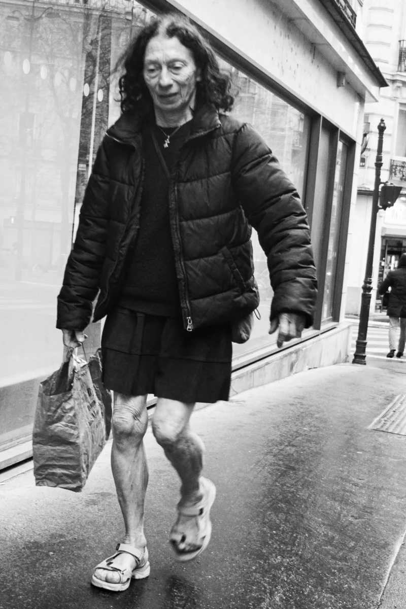 Alice · 11/04/2026 · 1649 x 2474 Cette photographie en noir et blanc met en scène une personne à l’allure décontractée, marchant en ville. Le sujet principal, une femme aux cheveux longs et vêtue d’une veste matelassée et d’une jupe courte, tient un sac en papier.La composition est simple et naturelle, avec un cadrage serré qui isole le sujet. La lumière semble douce et diffuse, typique d’un jour ordinaire. Les contrastes sont modérés, accentuant les détails du visage et des vêtements.L’ambiance est urbaine et intime, évoquant une promenade quotidienne. Le style est documentaire, réaliste et dépourvu d’artifice.