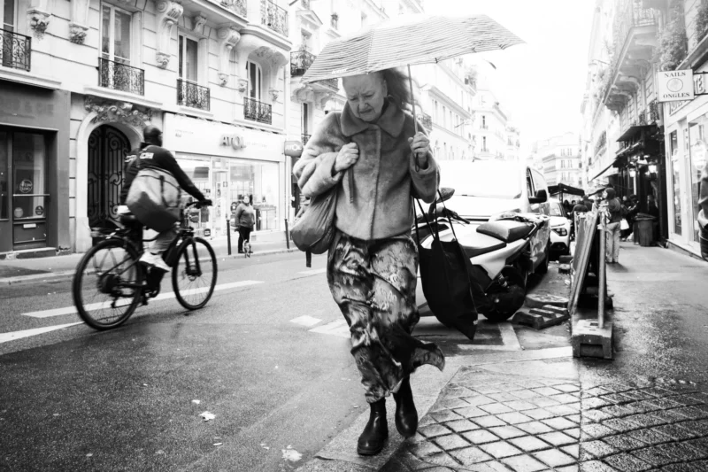 Alice · 11/04/2026 · 4734 x 3156 Cette photographie en noir et blanc met en scène un homme comme sujet principal, transportant un objet volumineux sous un parapluie. La composition montre une rue animée avec des cyclistes et des bâtiments en arrière-plan.Le cadrage est serré sur le sujet, tout en intégrant des éléments urbains. La lumière, douce et diffuse, crée des contrastes modérés. L’ambiance est dynamique et quotidienne, reflétant une vie urbaine trépidante. Le style est documentaire, capturant un instant authentique. Les tons de gris dominent, accentuant les textures et les mouvements.