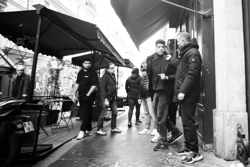 Alice · 11/04/2026 · 4133 x 2755 Cette photographie en noir et blanc capture un groupe de personnes en discussion dans une rue marchande. Le sujet principal semble être les quatre individus au centre, vêtus de manière décontractée.La composition montre une scène urbaine avec des étals couverts de marchandises sous des auvents. Le cadrage est serré, mettant en valeur les interactions humaines. La lumière est naturelle, mais contrastée, créant des ombres marquées. Les couleurs, bien que monochromes, soulignent les textures et les contrastes lumineux.Le style est documentaire, reflétant une ambiance authentique et quotidienne, presque intimiste malgré la présence de plusieurs personnes.